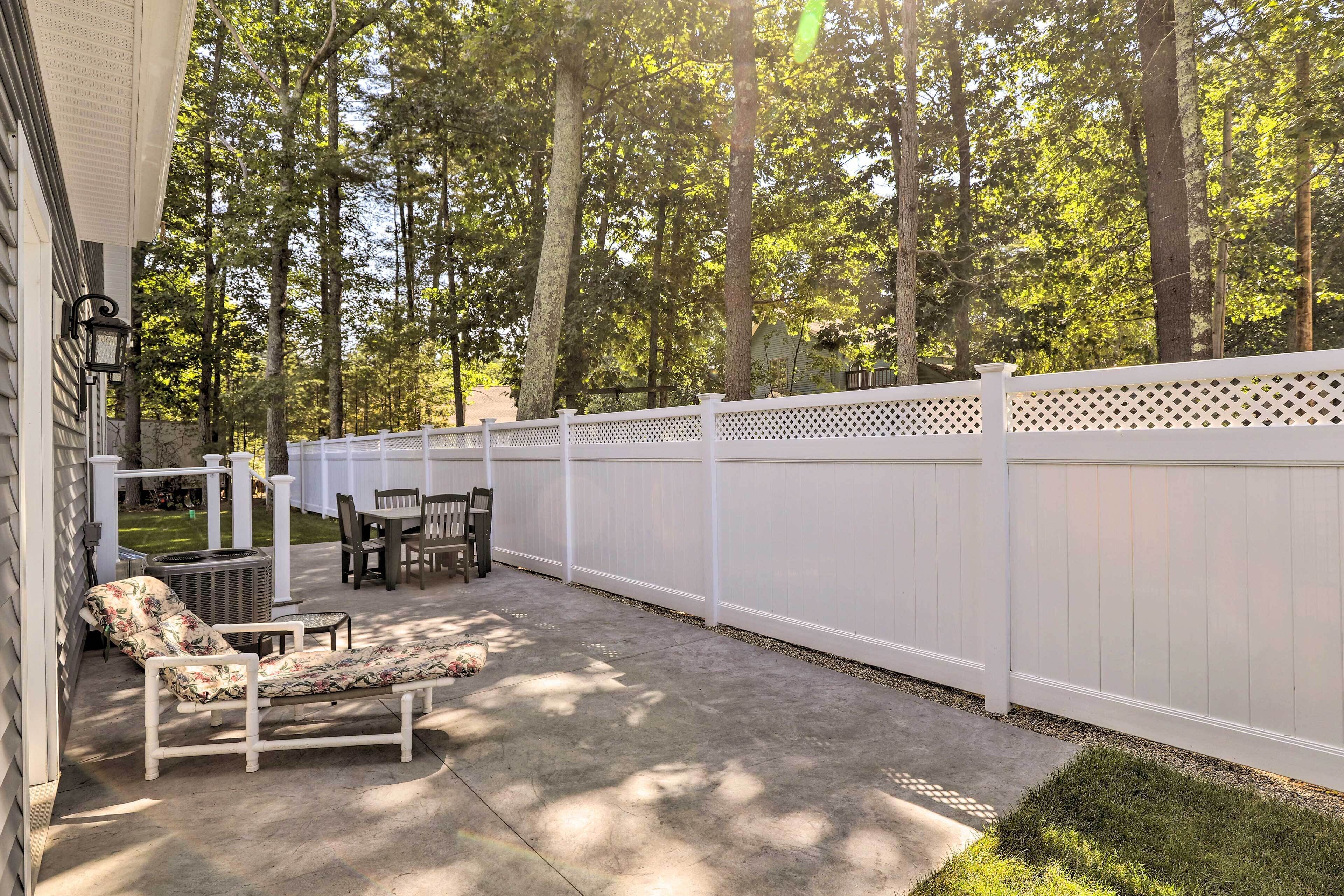 Un patio couvert, délimité par une clôture blanche, agrémenté de mobilier d’extérieur et offrant une vue sur une zone boisée.