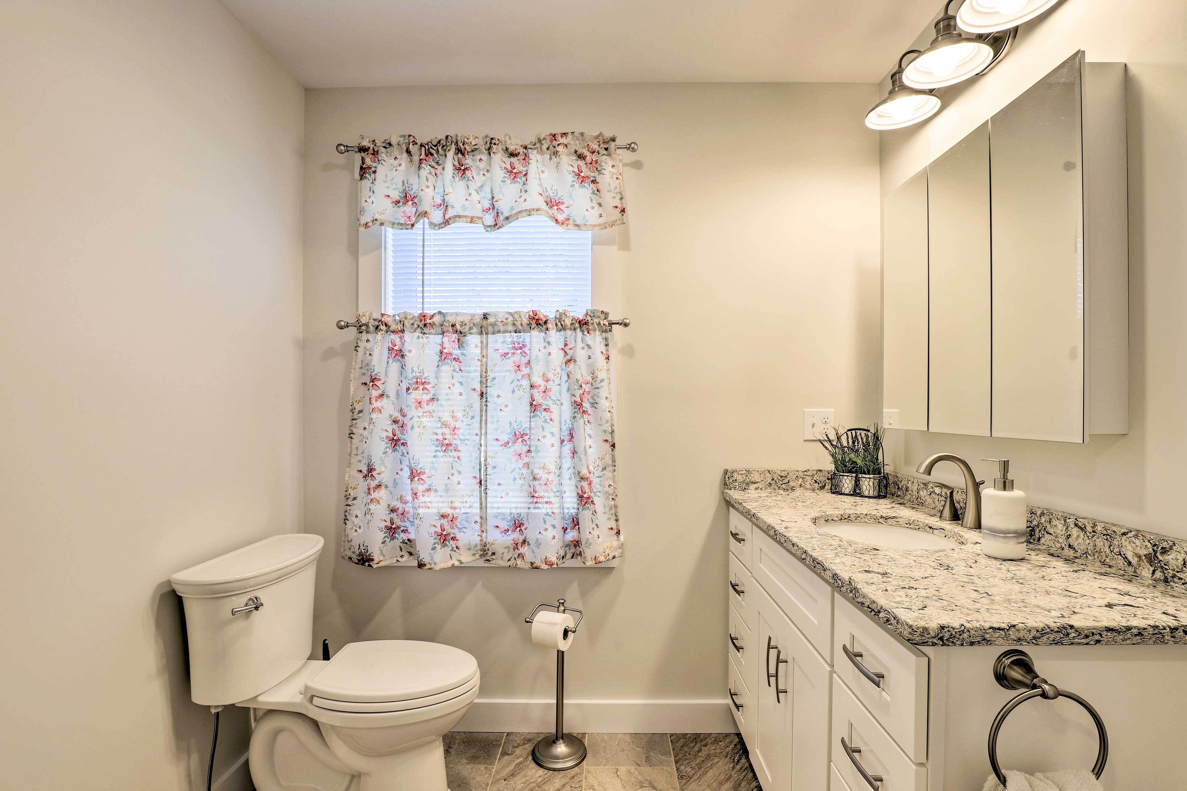 Une salle de bain comprenant des toilettes, un lavabo avec un plan de toilette en marbre, un miroir et une fenêtre agrémentée de rideaux à motifs floraux.