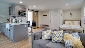 Ett kompakt bostadsutrymme med ett litet kök, en soffa med mönstrade kuddar, en säng med dekorativa prydnadskuddar och en träbänk.