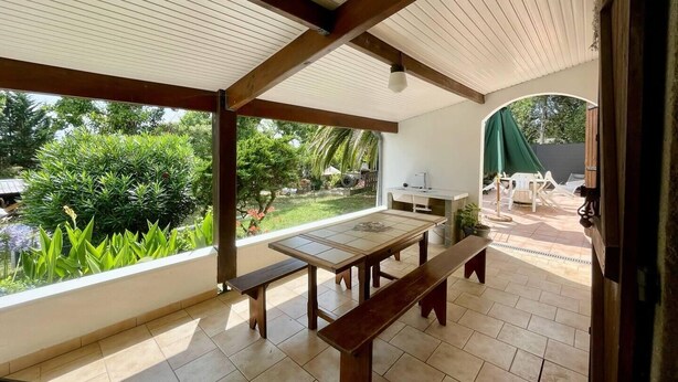 A covered patio with a dining table and benches, a sink, and a view of a garden.