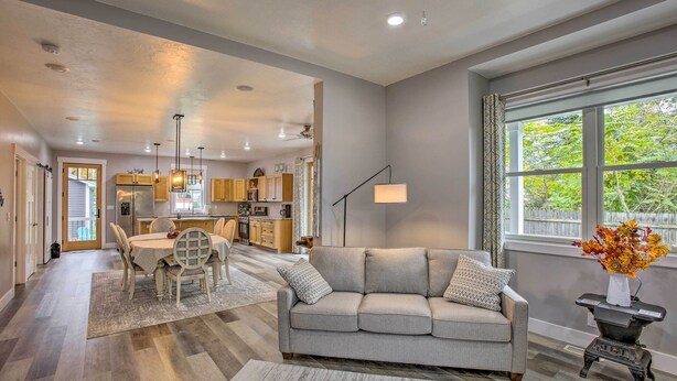 A modern living room with a grey sofa, dining table, and chairs. There is a kitchen in the background with wooden cabinets and a window with a view of trees. - < 1 Mi to Dtwn Ludington: Bright Lower-level Unit (Ludington)