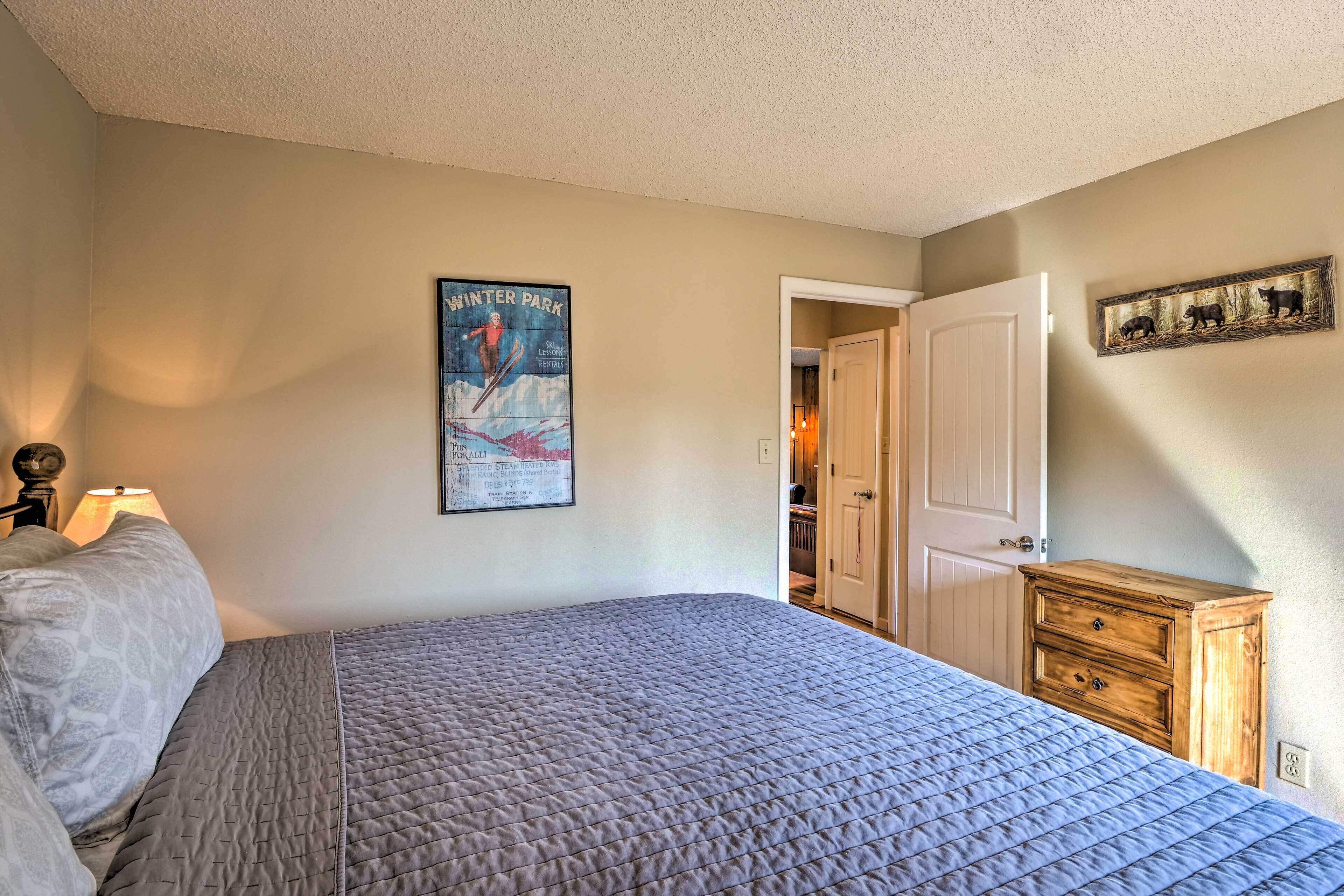 Un dormitorio con cama, mesita de noche, cómoda, un cartel en la pared y una puerta que da a otra habitación.