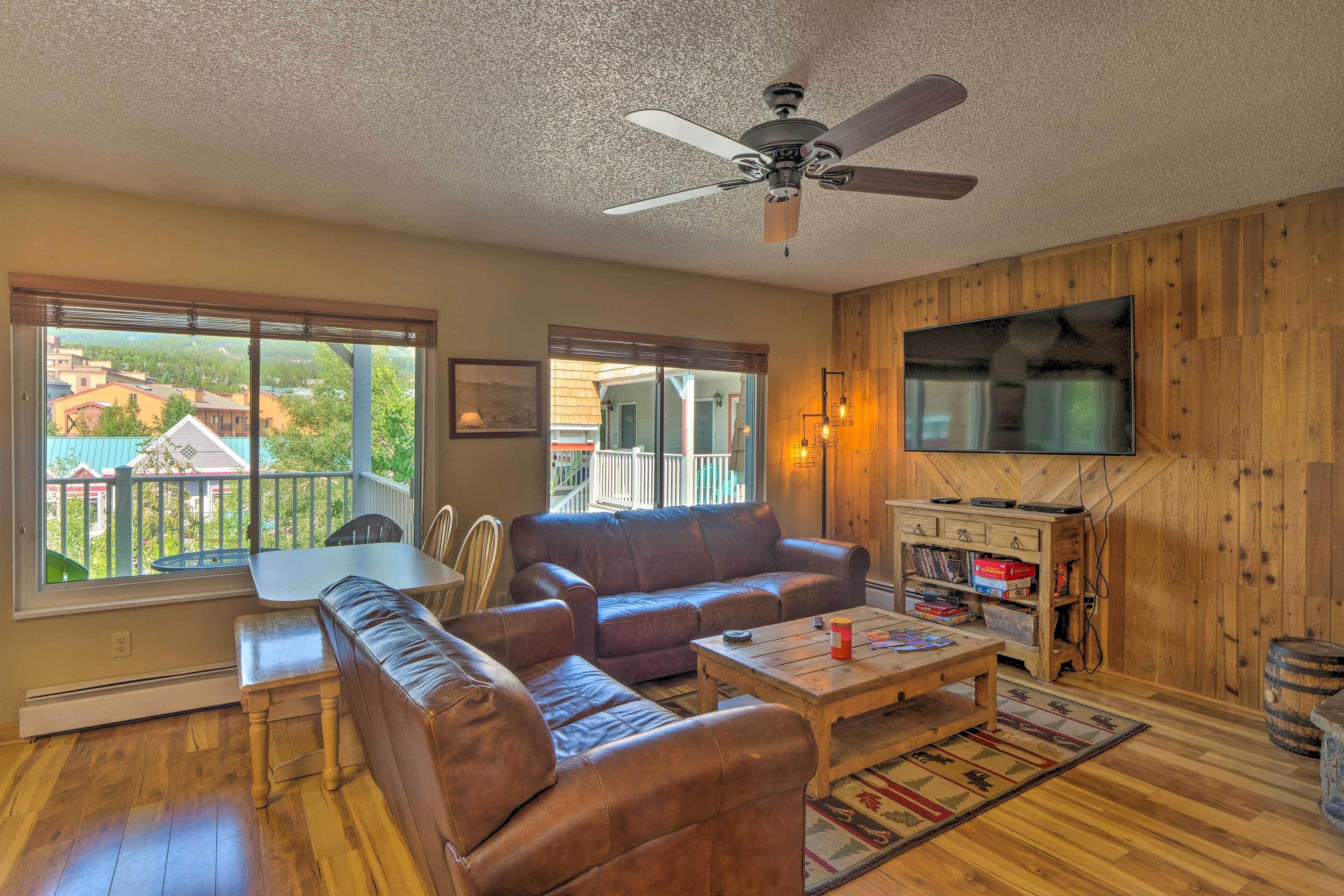 Ein gemütliches Wohnzimmer mit Holzboden, Deckenventilator, großem Flachbildfernseher, Couchtisch aus Holz, braunem Ledersofa, einer Bank und Blick auf einen Balkon mit Gartenmöbeln.