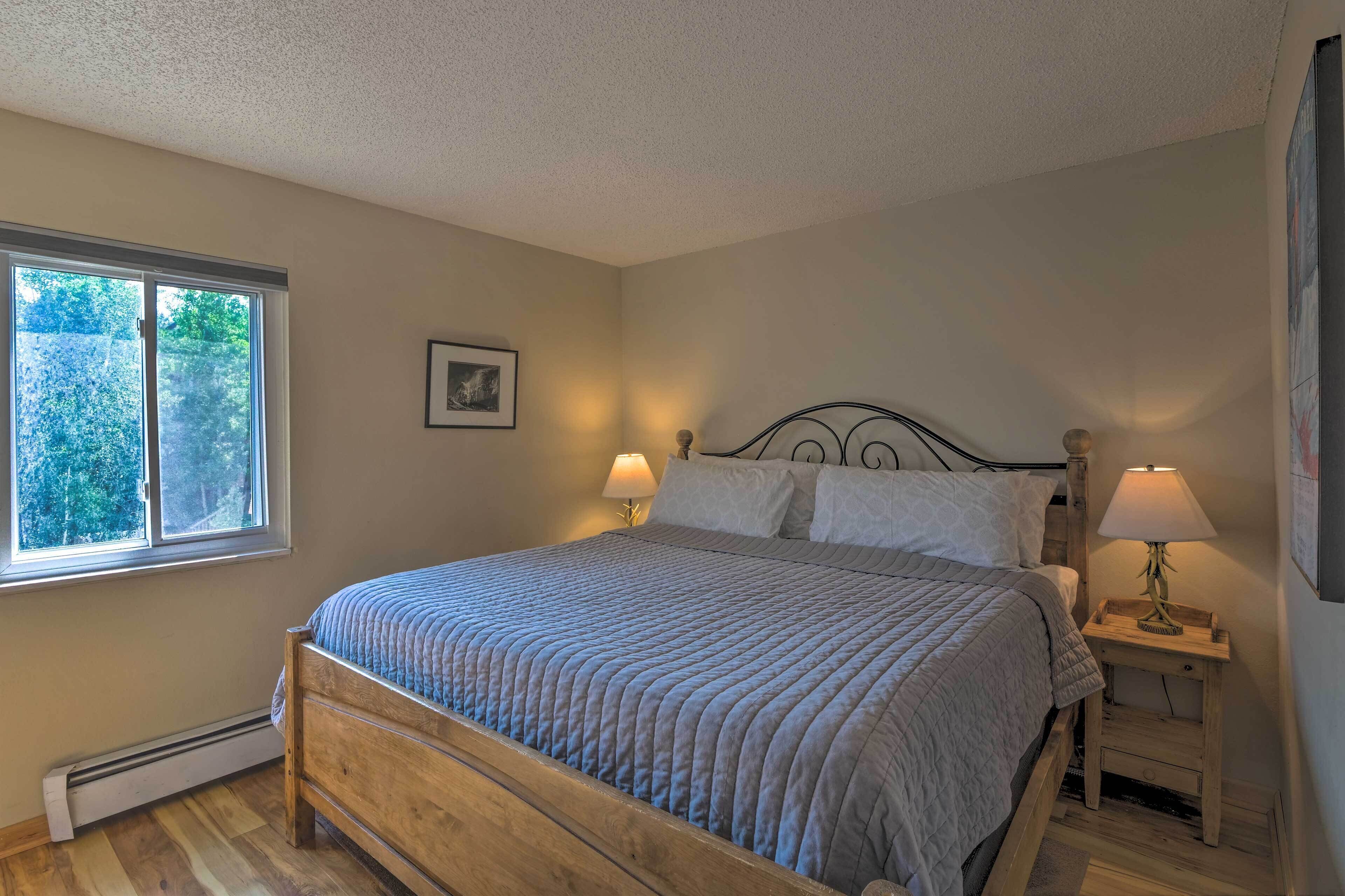A bedroom with a wooden bed, a window, a nightstand with a lamp, and a wall-mounted picture.