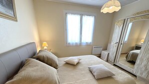 Une chambre à coucher avec un lit, des oreillers, une table de chevet, une lampe, une fenêtre, un radiateur et un miroir.
