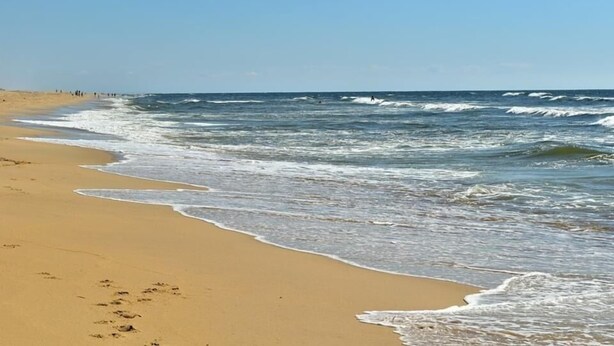 Une plage où les vagues viennent doucement lécher le rivage, un ciel dégagé et quelques personnes aperçues au loin.