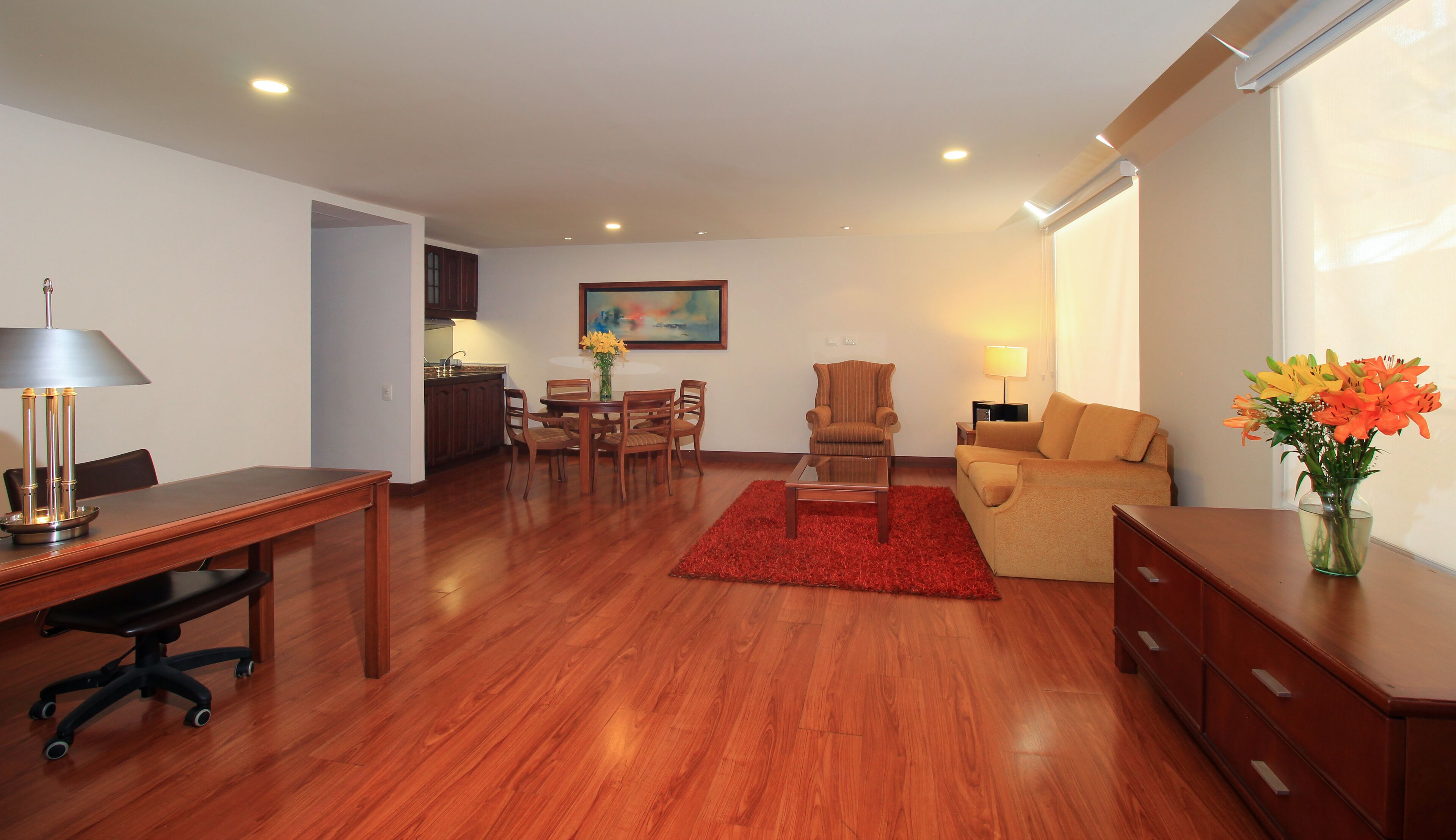 A spacious living room with a wooden floor, a dining area with a table and chairs, a sofa, a red rug, and a vase with flowers.