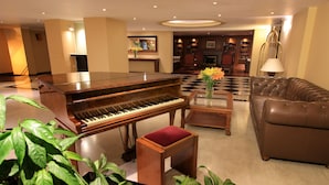 A grand piano, a leather sofa, a coffee table, and potted plants in a lobby area. - Hotel Windsor House Inn By GEH Suites (Bogotá)