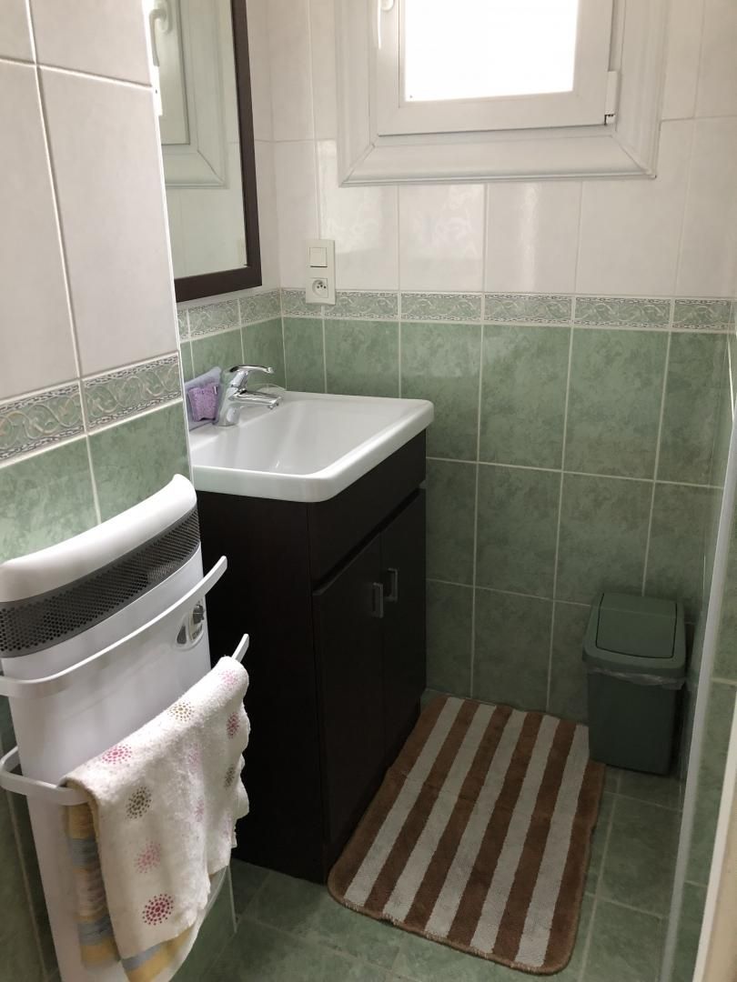 A small bathroom with a white sink, dark cabinets, a green tiled wall, a window, and a striped rug.