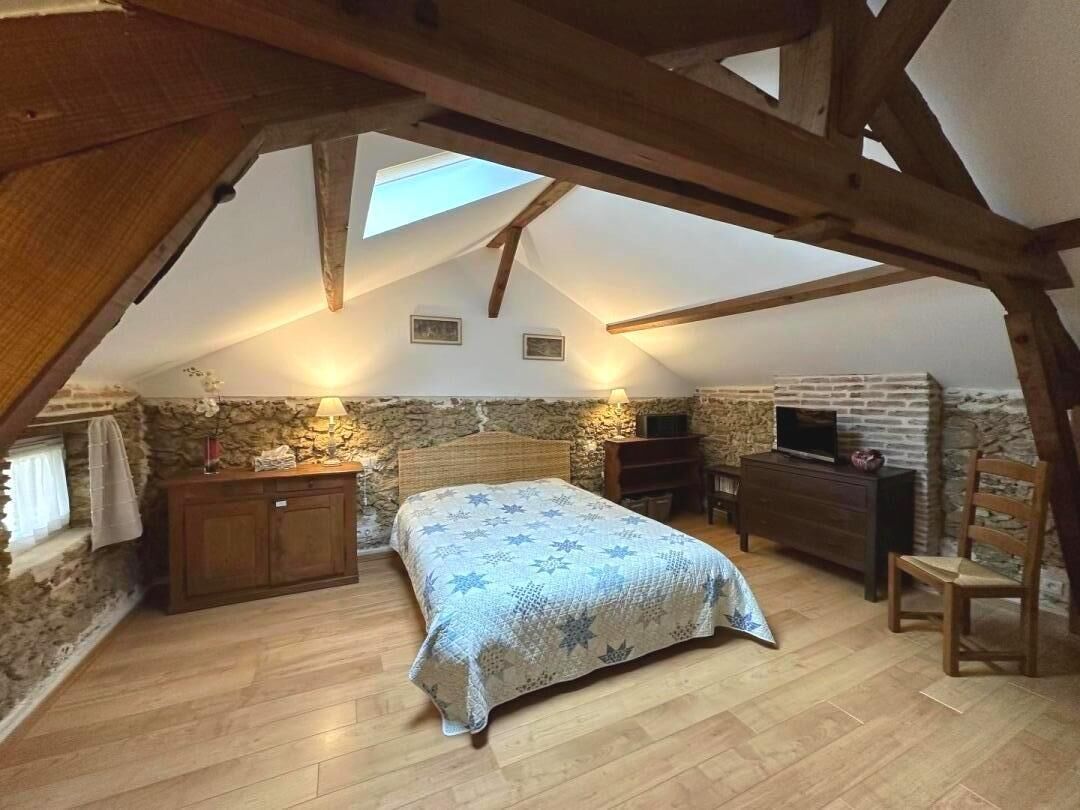 A bedroom with a sloped ceiling, stone walls, a bed with a patterned bedspread, a wooden dresser, a chair, and a television.