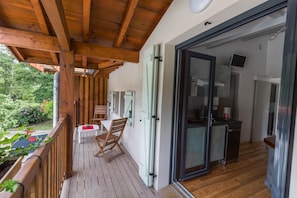 Um deck de madeira com mesa e cadeiras, uma porta de correr envidraçada que dá para uma cozinha e vista para o verde lá fora.