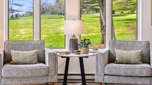 A room with two grey upholstered chairs, a small table with a lamp and decorative items, and large windows offering a view of a grassy landscape. - Sauna Condo with Golf Views, Coach Road (Quechee)