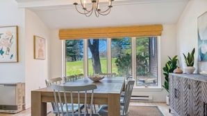 A dining area with a wooden table, grey chairs, a chandelier, and a view of a garden through large windows. - Sauna Condo with Golf Views, Coach Road (Quechee)