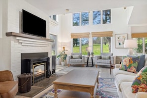 A spacious living room with a fireplace, a television, and a large window overlooking a garden. - Beautiful Golf Course Views, 3BR - Coach Road 13B (Quechee)