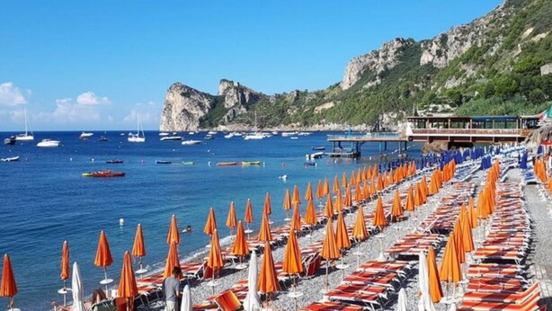 A beach with rows of orange umbrellas and lounge chairs, a rocky coastline, and boats in the water. - New apartment 100 meters from the sea (Nerano)