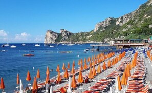 Ein Strand mit Reihen orangener Sonnenschirme und Liegestühle, eine felsige Küstenlinie und Boote im Wasser.