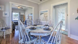 Une salle à manger avec une table ronde, des chaises blanches et un lustre. Les murs sont ornés de tableaux encadrés et il y a une fenêtre avec des rideaux.