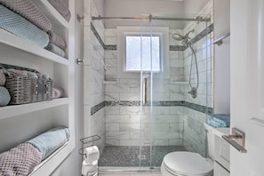 Une salle de bain avec une douche à parois de verre, des toilettes et des étagères où sont pliées des serviettes.