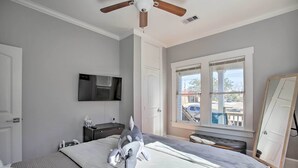 Une chambre à coucher avec un lit, un ventilateur de plafond, un téléviseur fixé au mur et une fenêtre avec des stores.