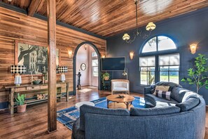 A cozy living room with a wooden ceiling, a large sofa, a coffee table, and a TV. There are two archways leading to another room, and a large window with a view. - Stunning Historic Home < 4 Mi to Beaches! (Galveston)