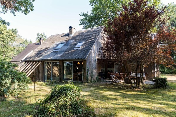 Een rustiek houten huis met een schuin dak, grote ramen en een patio met buitenmeubels.