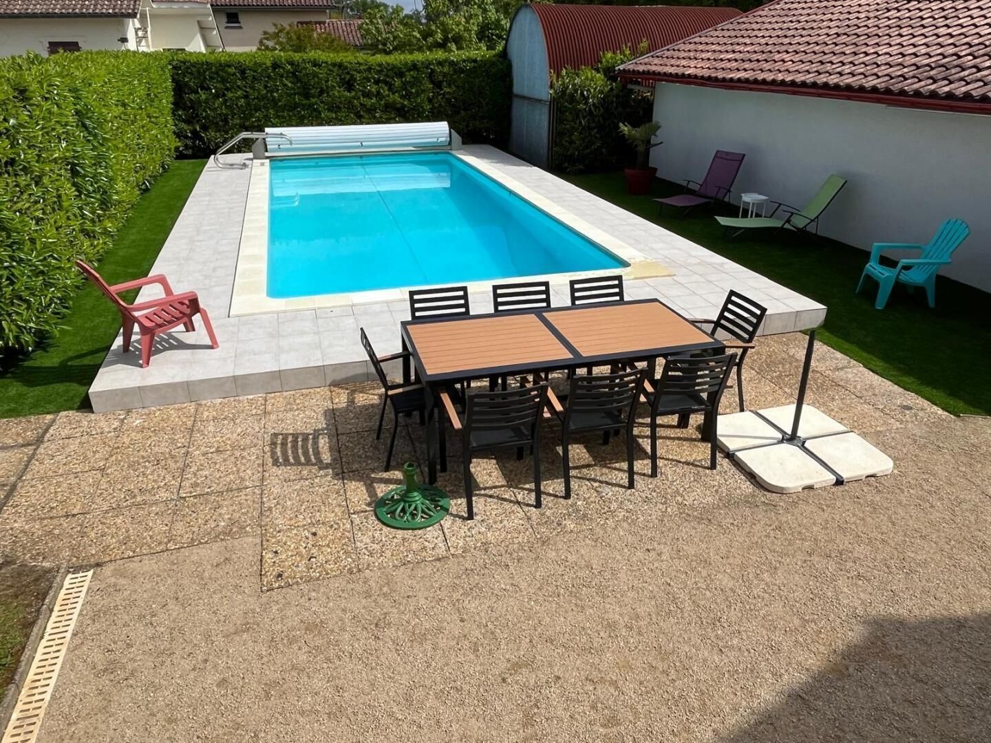A swimming pool with a tiled deck, surrounded by a patio area with a table and chairs, and a small garden with a hedge and chairs.