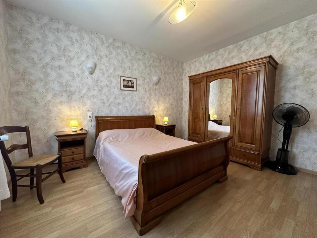 Um quarto com cama de madeira, guarda-roupa de madeira, uma cadeira, mesa de cabeceira com abajur e um ventilador.