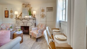 Una sala de estar acogedora con chimenea de piedra, sillones estampados, una mesa de comedor preparada para cuatro personas y un gran ventanal que proporciona luz natural.