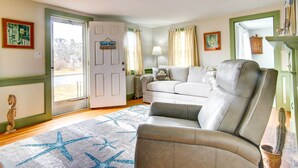 Un salon avec un fauteuil beige, un canapé blanc, un tapis à motifs d’étoiles, une porte portant l’inscription « Beach House » et un cactus dans un pot.