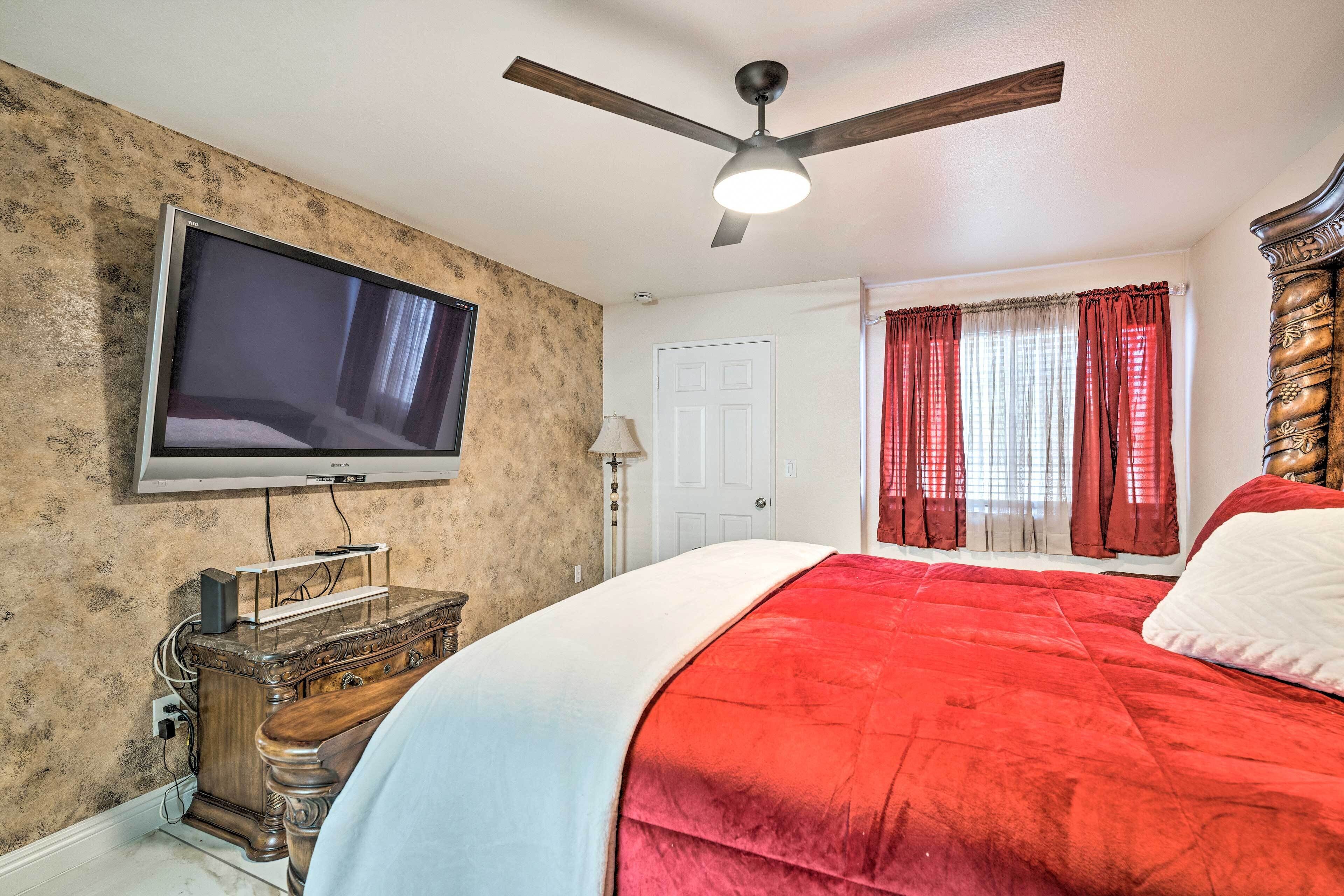 Ein Schlafzimmer mit einem großen Bett, einem Deckenventilator, einem an der Wand montierten Fernseher und einem Fenster mit roten Vorhängen.
