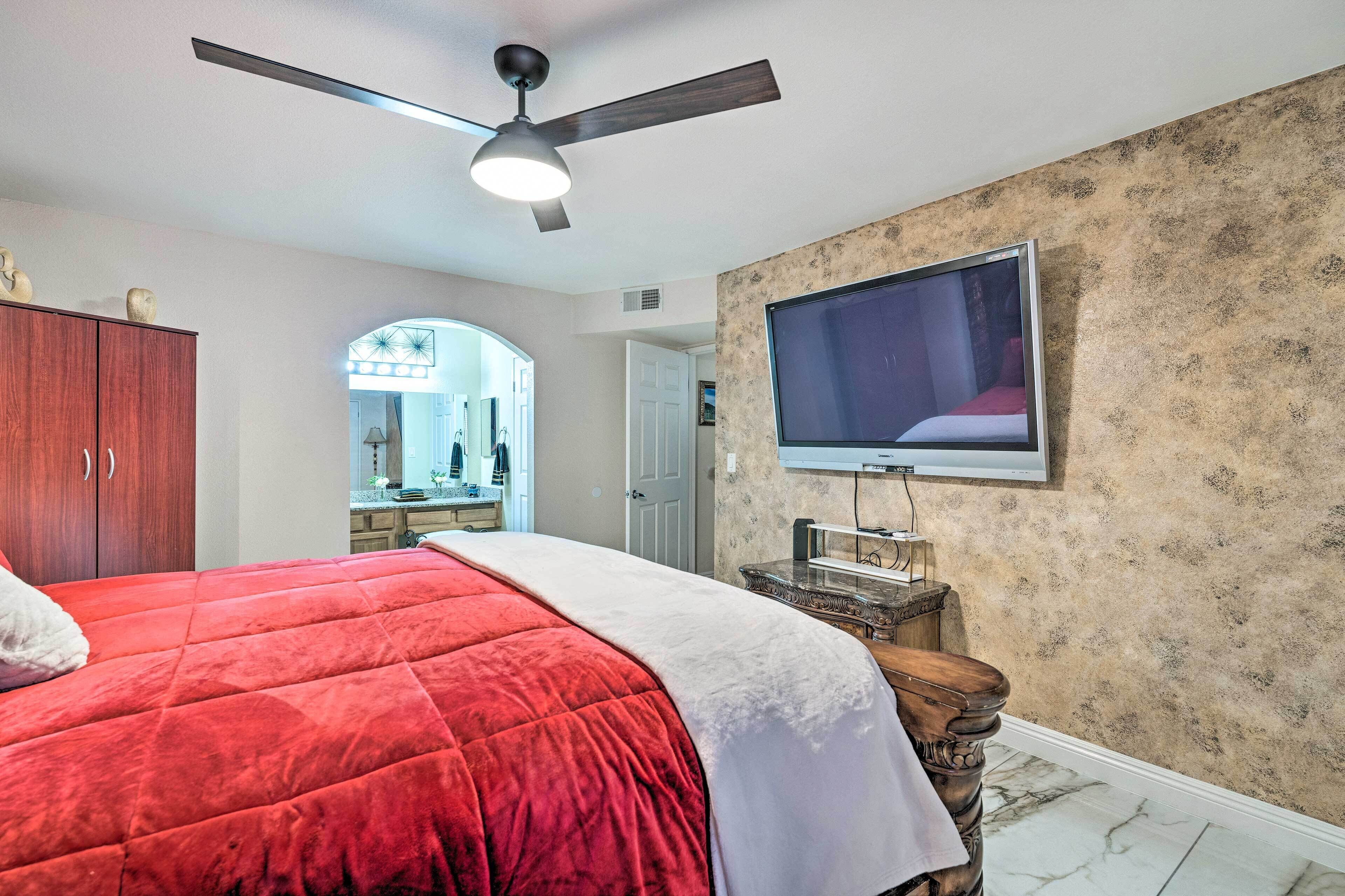 A bedroom with a large bed, a ceiling fan, a TV mounted on the wall, and a wooden nightstand.