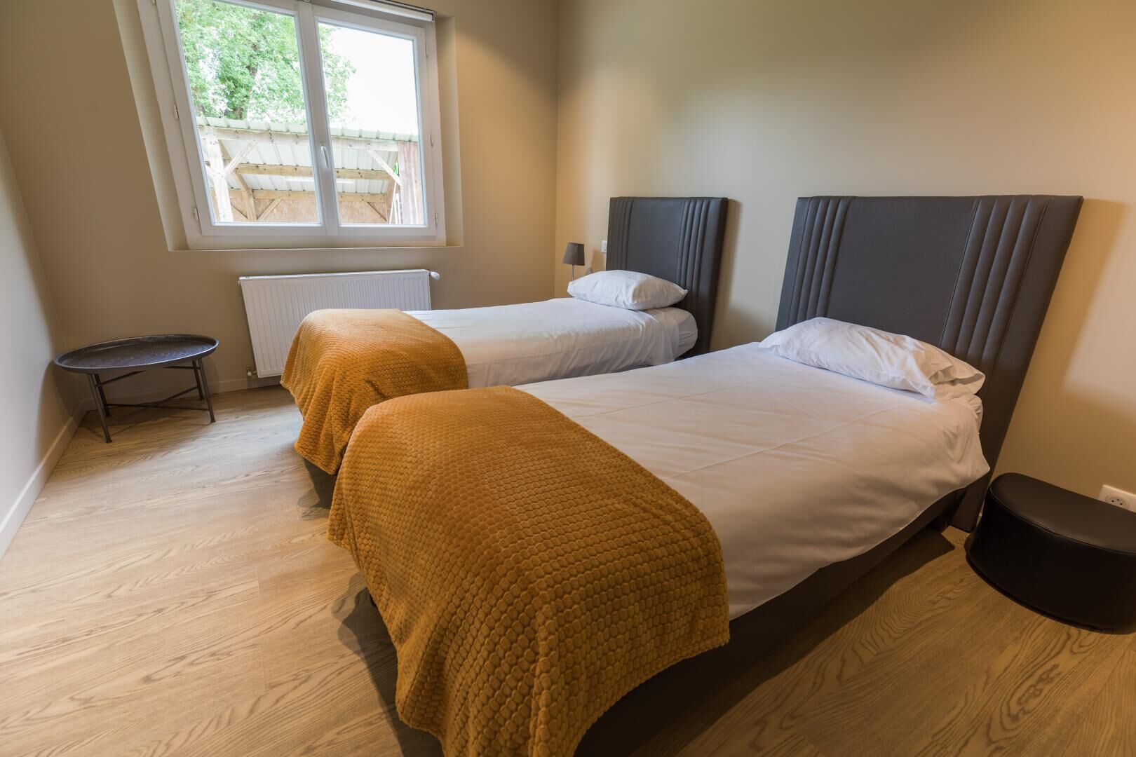 A hotel room with two beds, a small table, a radiator, and a window with a view of the outside.