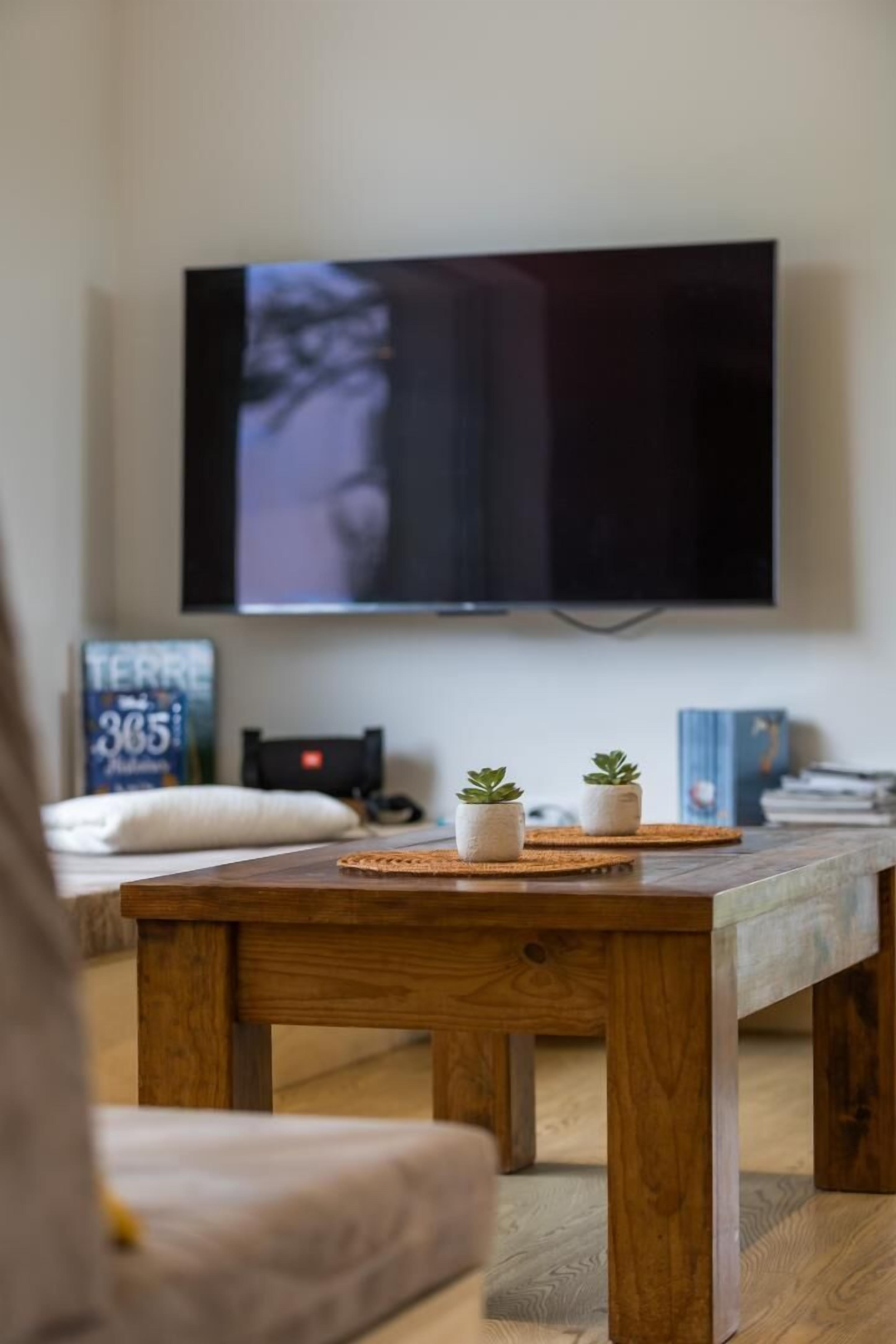 Ein Wohnzimmer mit Couchtisch aus Holz, zwei Topfpflanzen, einem flachbildfernseher an der Wand und einem Bücherregal mit Büchern und einer Uhr.