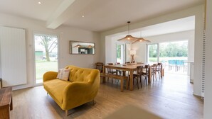 A spacious living area with a yellow sofa, wooden dining table, and chairs. There are large windows providing natural light and a view of the outdoors. - Villa Cassora - Herm (HERM)