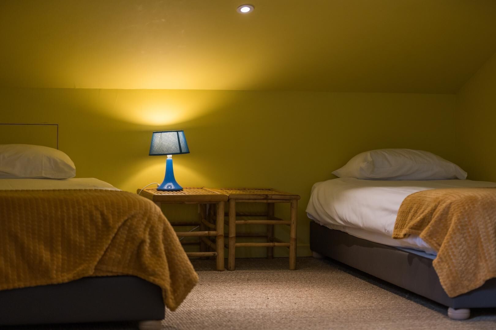 A small, dimly lit room with two beds, a small table, and a single lamp.