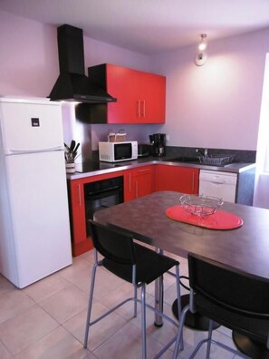 Cocina compacta con armarios rojos, un refrigerador blanco, microondas y cafetera. Hay una mesa de comedor con dos sillas y un mantel individual rojo.