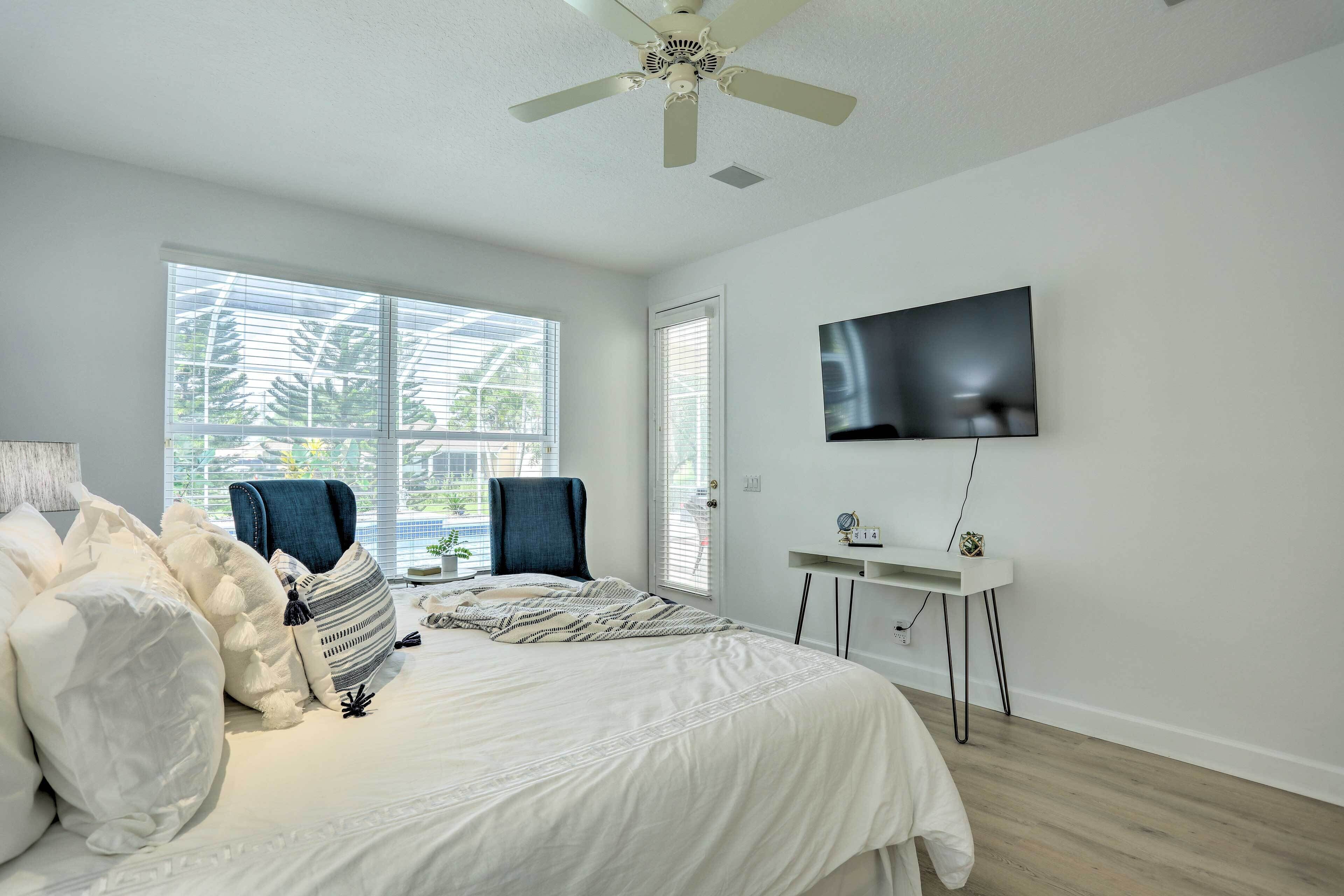 A bedroom with a bed, a desk, a chair, a TV, and large windows.