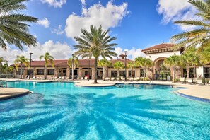 A large outdoor swimming pool with palm trees and lounge chairs, surrounded by a building with a tiled roof.
