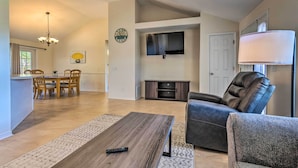 Un salón espacioso con una mesa de centro de madera, un sofá de cuero, un televisor montado en la pared y una zona de comedor con sillas y una mesa de madera.