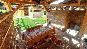 A wooden structure with a table covered in a quilt, a hanging light fixture, and a view of a fenced backyard with a barbecue area. - Cozy Alpine Apartment near Lake Bohinj,  Free Bikes & No Extra Fees (Bohinjska Bistrica)