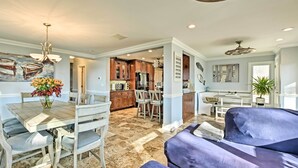 A spacious living area with a dining table, chairs, and a blue sofa. A kitchen is visible in the background with wooden cabinets and a refrigerator.