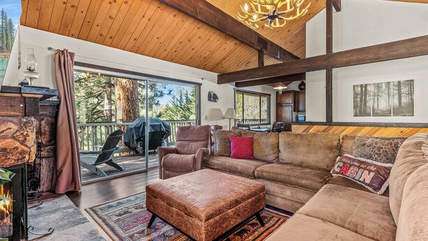 A cozy cabin interior with a fireplace, a large sofa, a cushioned ottoman, and a view of the outdoors. - 1 Mi to Slopes: Big Bear Ski Haven w/ Hot Tub (Big Bear Lake)