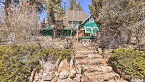 A rustic lodge with a stone staircase leading to a wooden deck, surrounded by dense trees and shrubs. - < 2 Mi to Lake: Big Bear City Cabin w/ Hot Tub! (Big Bear City)