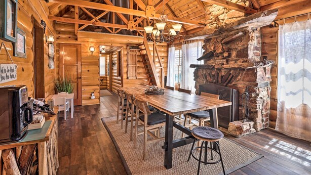 A rustic cabin interior with a wooden dining table, chairs, a fireplace, and a staircase leading to another room. - < 2 Mi to Lake: Big Bear City Cabin w/ Hot Tub! (Big Bear City)