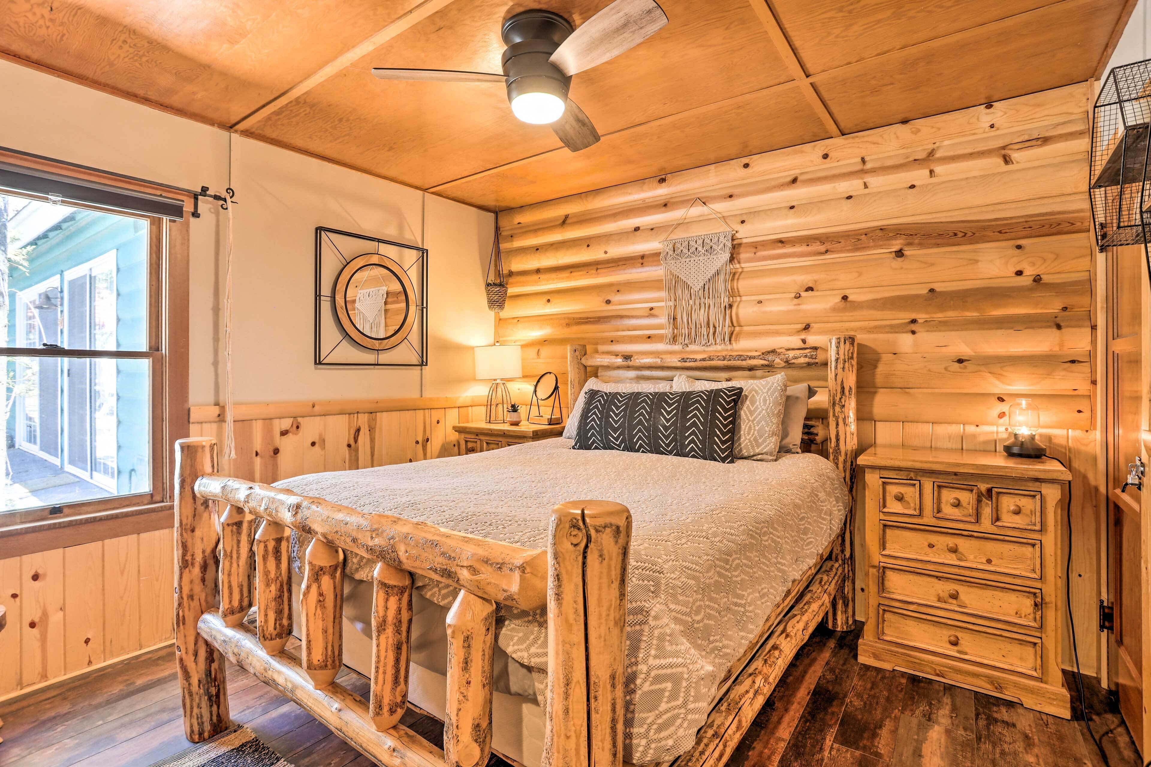 A log cabin-style bedroom with a wooden bed frame, a ceiling fan, a bedside table with a lamp, and a window with curtains.