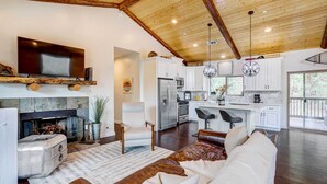 A spacious living room with a fireplace, a large TV mounted on a wooden beam, and a cozy seating area with a leather couch and a coffee table. - Custom-built Gem w/ Kayaks & Games in Big Bear! (Big Bear City)