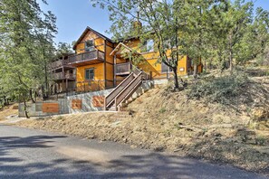 A multi-story wooden cabin with a balcony and stairs leading to the upper floor, surrounded by trees and a dirt slope. - Custom-built Gem w/ Kayaks & Games in Big Bear! (Big Bear City)