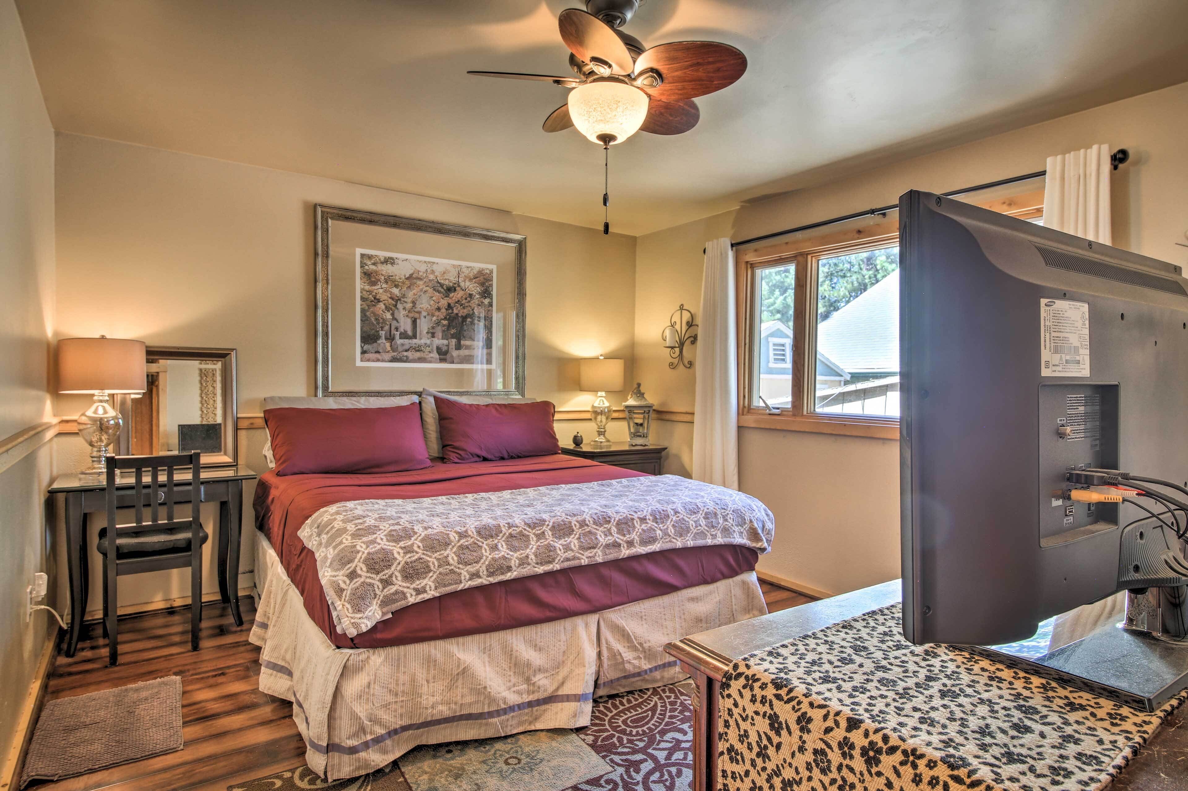 A bedroom with a bed, desk, chair, ceiling fan, and a television.
