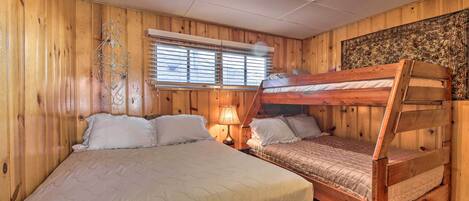Una habitación de madera con una cama grande, otra más pequeña y una cama nido. Hay una ventana con persianas y un adorno metálico en la pared.