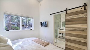 A bedroom with a bed, a window with a view of trees, a TV, and a sliding door with a wooden panel design. - Half-mi to Big Bear Lake: Modern Home w/ Deck (Big Bear Lake)